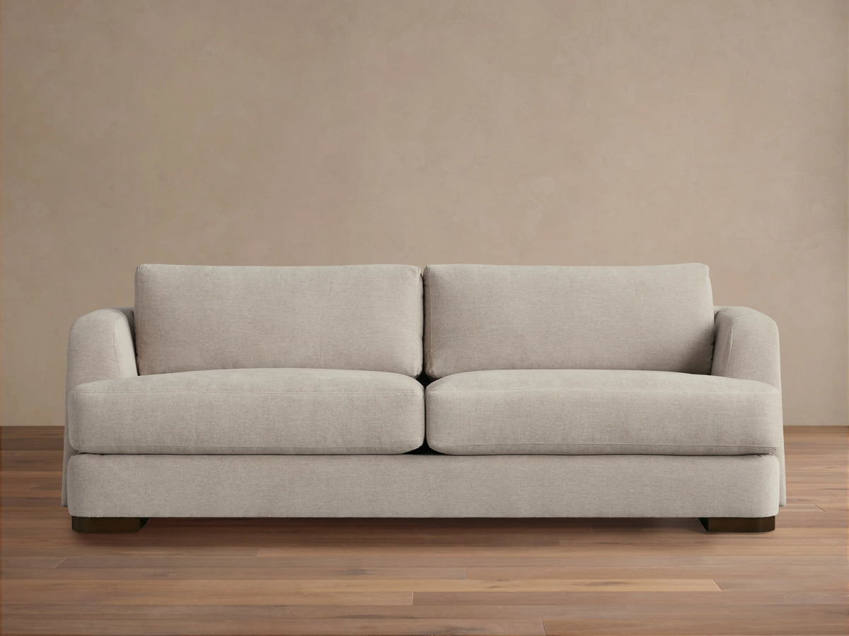 A beige Kayden Sofa with plush seat and back cushions, featuring a simple, modern design. Set on a wooden floor against a neutral wall.