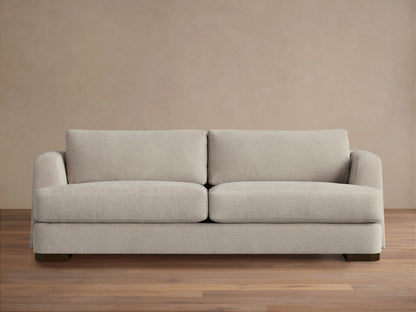 A beige Kayden Sofa with plush seat and back cushions, featuring a simple, modern design. Set on a wooden floor against a neutral wall.