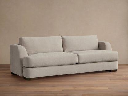 A beige Kayden Sofa with plush seat and back cushions, featuring a simple, modern design. Set on a wooden floor against a neutral wall.