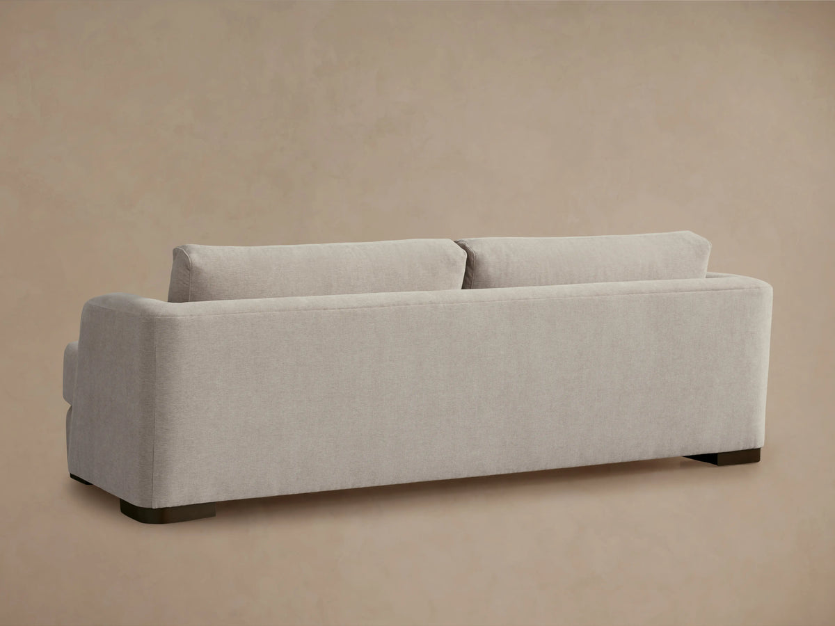 Alt text: The Kayden Sofa in a light beige fabric, featuring a rounded armrest and plush back cushions. The sofa has a smooth texture and sturdy wood base.
