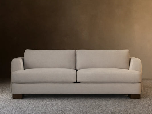 A beige Kayden Sofa with plush seat and back cushions, featuring a simple, modern design. Set on a wooden floor against a neutral wall.