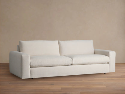 Kipton Wide Arm Sofa, two-seater, in textured off-white fabric, with wide squared arms. Placed on a wood floor against a tan wall.
