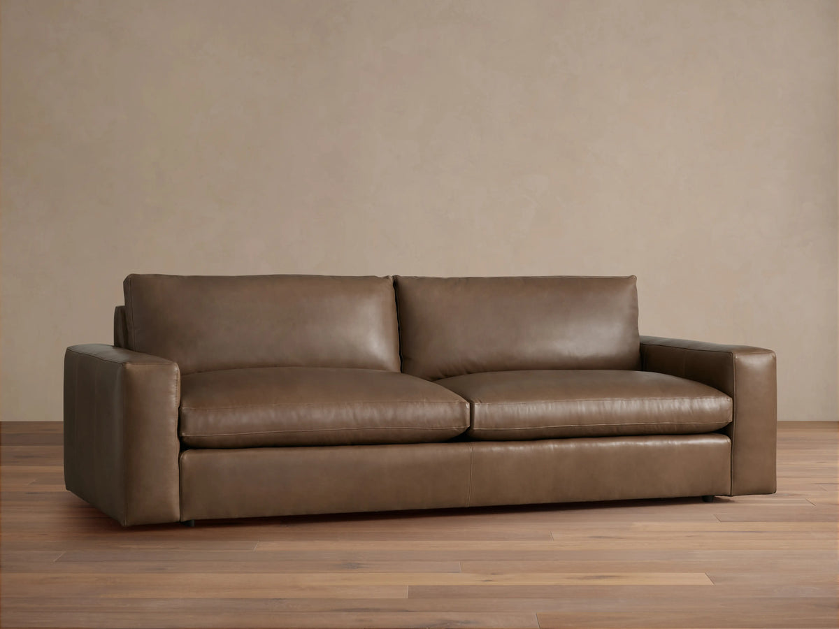 Kipton Wide Arm Leather Sofa. Warm brown leather, wide square arms, two plush seat & back cushions, on a wood floor.