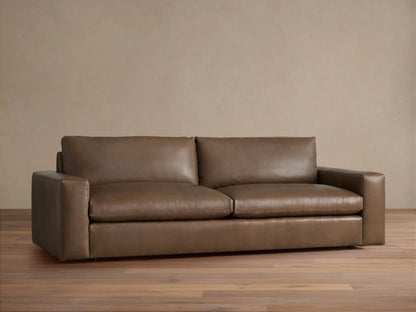 Kipton Wide Arm Leather Sofa. Warm brown leather, wide square arms, two plush seat & back cushions, on a wood floor.