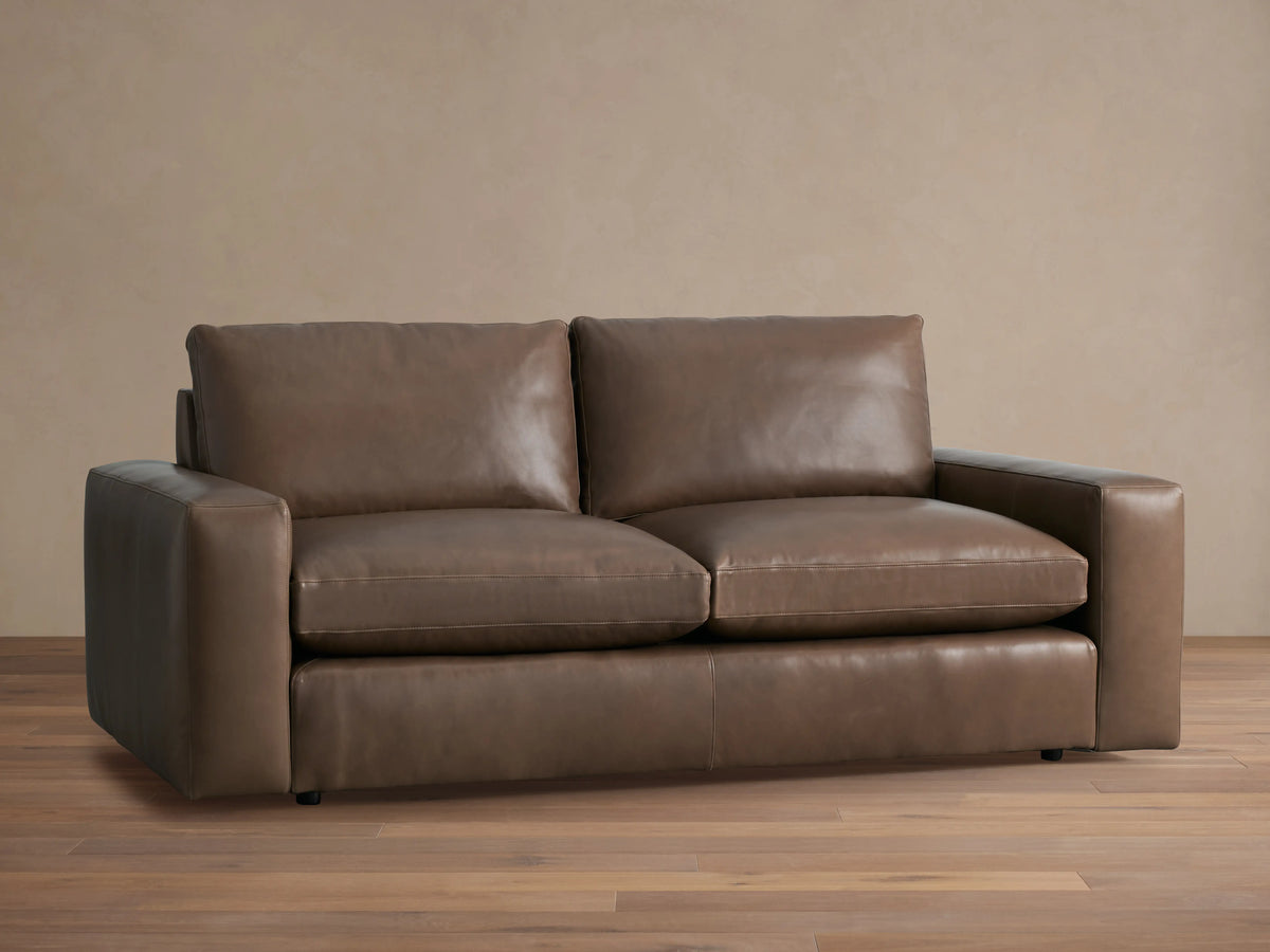 Kipton Wide Arm Leather Sofa, two-seater, in warm brown leather, with plush cushions, on a wooden floor against a neutral wall.