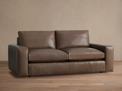 Kipton Wide Arm Leather Sofa, two-seater, in warm brown leather, with plush cushions, on a wooden floor against a neutral wall.
