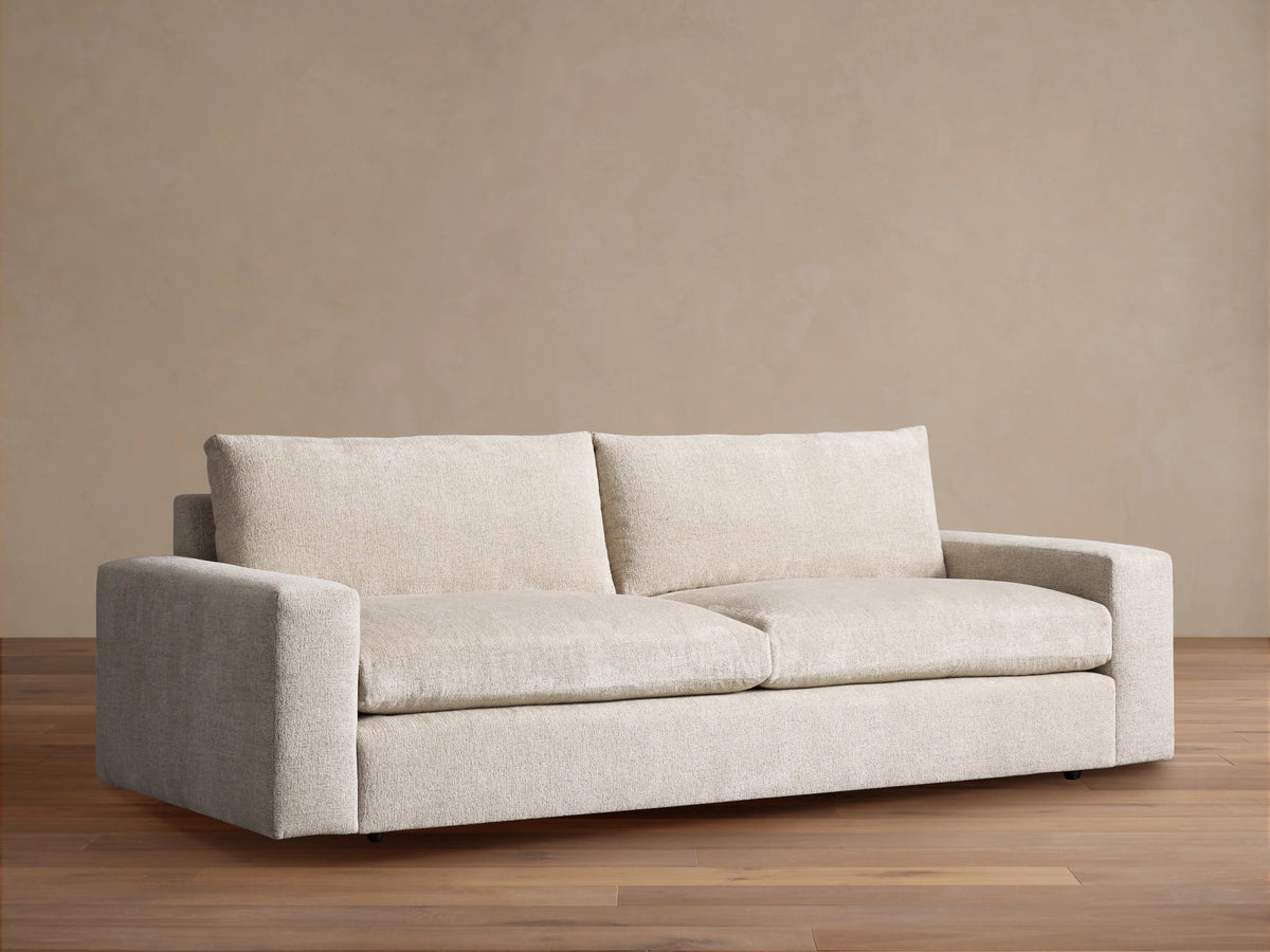 Light cream Kipton Wide Arm Sofa with two seat and back cushions, on a wood floor against a beige wall.