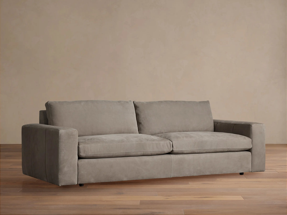 Kipton wide arm nubuck leather sofa in light taupe, with two seat/back cushions, on wood floor, against a beige wall.