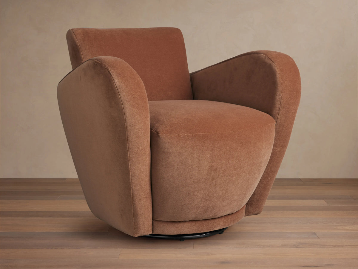 Front-right view of a plush, curved, rust-colored velvet Amelia Swivel Chair on a black swivel base, set on a wooden floor.