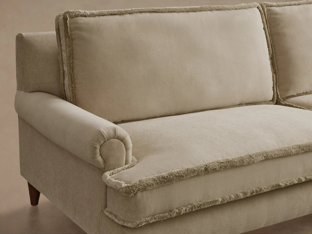 Front-side view of the beige McKinley Sofa, showing its rolled arm, fringed cushion edges, and a dark wooden leg.