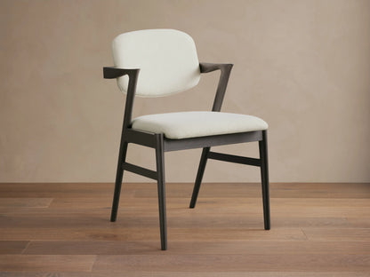 Marais Dining Arm Chair, dark ash wood frame with curved armrests, cream padded seat and back, angled on wood floor.