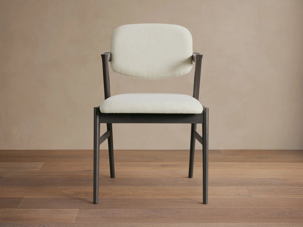 Marais Dining Arm Chair, dark ash wood frame with curved armrests, cream padded seat and back, angled on wood floor.