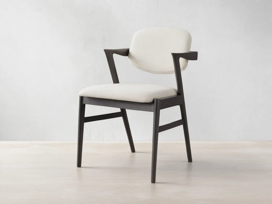 Marais Dining Arm Chair, dark ash wood frame with curved armrests, cream padded seat and back, angled on wood floor.