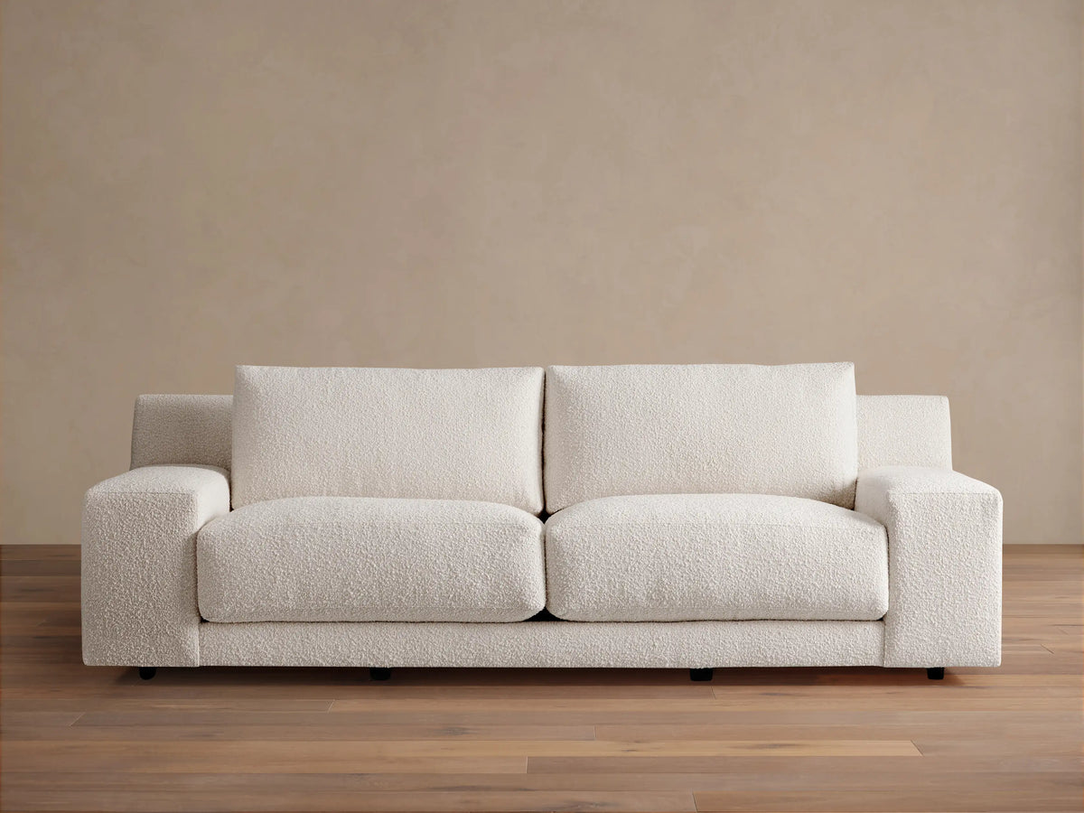 Novoli Sofa, cream boucle fabric, two seat cushions, square arms. Front view on a wood floor against a beige wall.