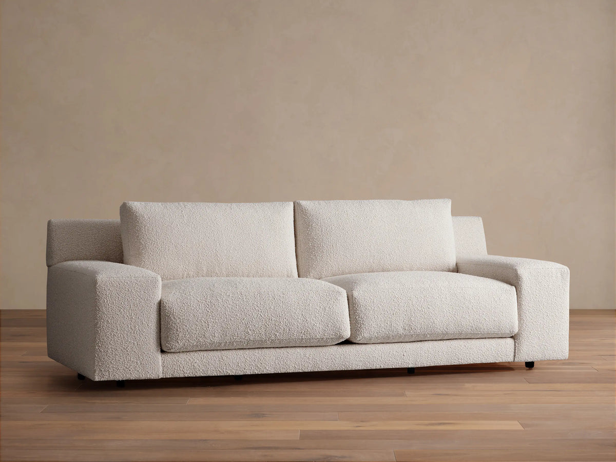 Novoli Sofa, cream boucle fabric, two seat cushions, square arms. Front view on a wood floor against a beige wall.