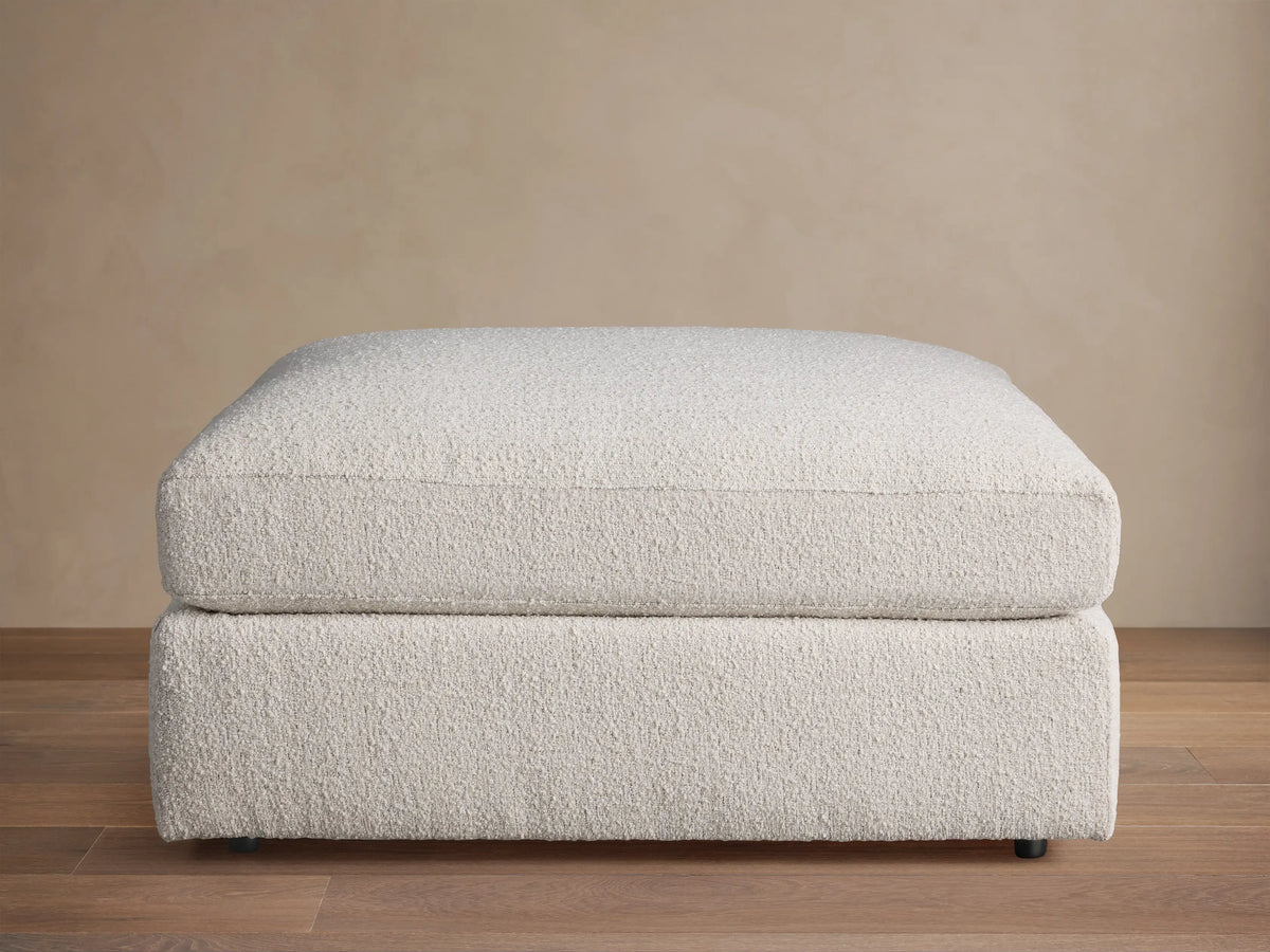 Novoli Ottoman, off-white textured fabric, rectangular, cushioned top on base, against a neutral wall and wood floor.