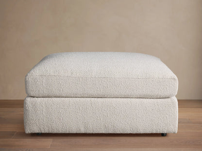 Novoli Ottoman, off-white textured fabric, rectangular, cushioned top on base, against a neutral wall and wood floor.