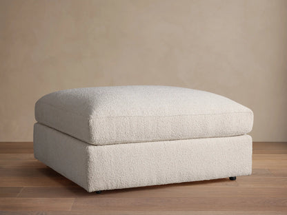 Novoli Ottoman, off-white textured fabric, rectangular, cushioned top on base, against a neutral wall and wood floor.