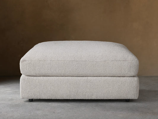Novoli Ottoman, off-white textured fabric, rectangular, cushioned top on base, against a neutral wall and wood floor.
