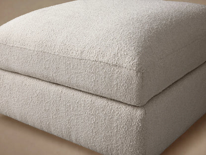 Novoli Ottoman in textured off-white performance fabric, featuring a soft, rounded top cushion over a rectangular base on a wood floor.