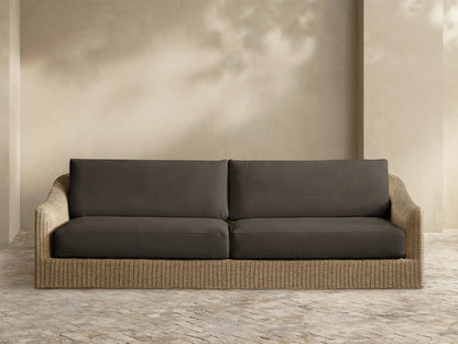 Front view of Ancona Outdoor Sofa, featuring a beige handwoven wicker frame and dark grey seat and back cushions on a stone patio.