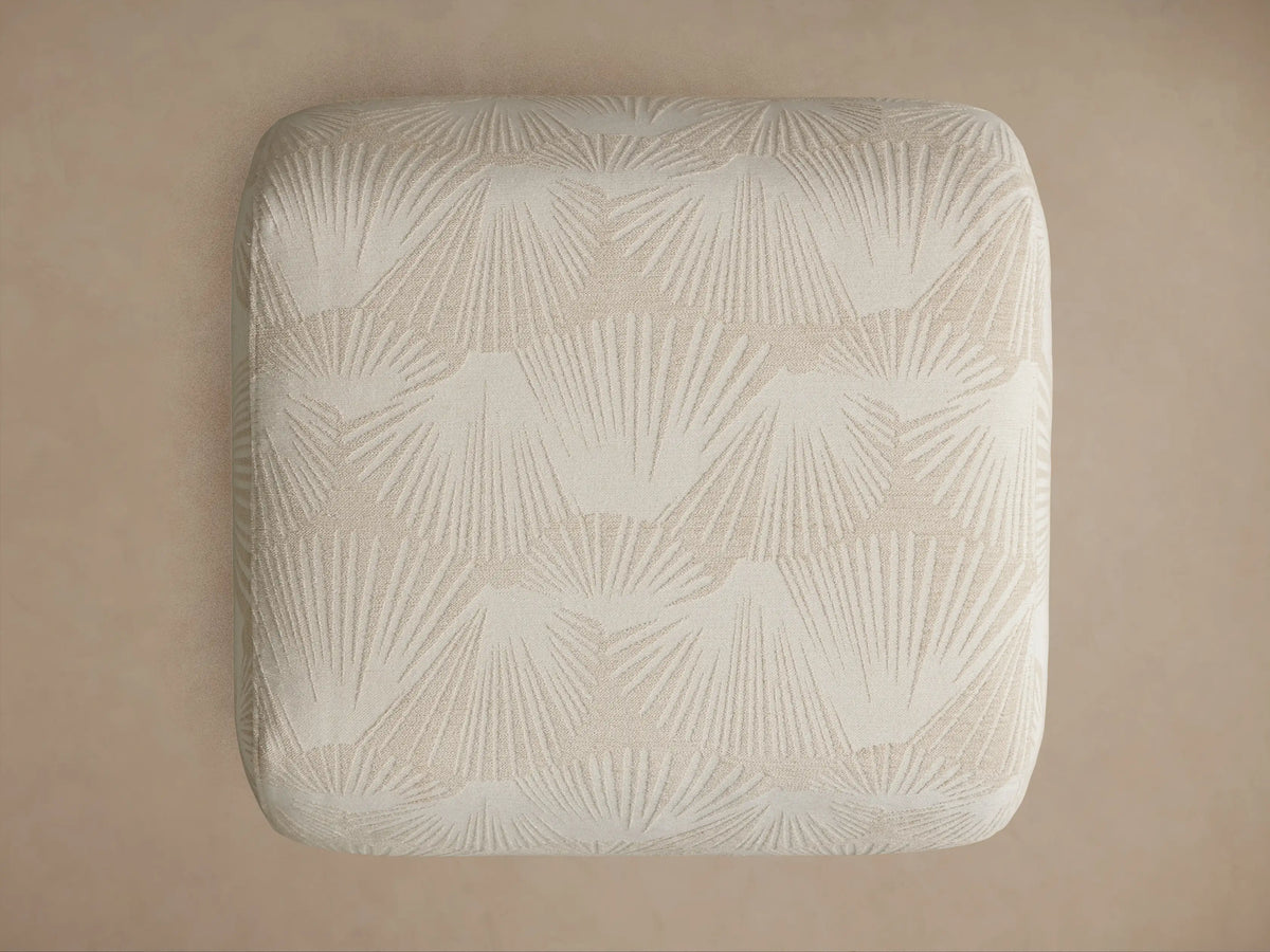 Close-up of a Fresno Ottoman, showing its light beige fabric with a subtle raised palm frond pattern on its corner and curved side.