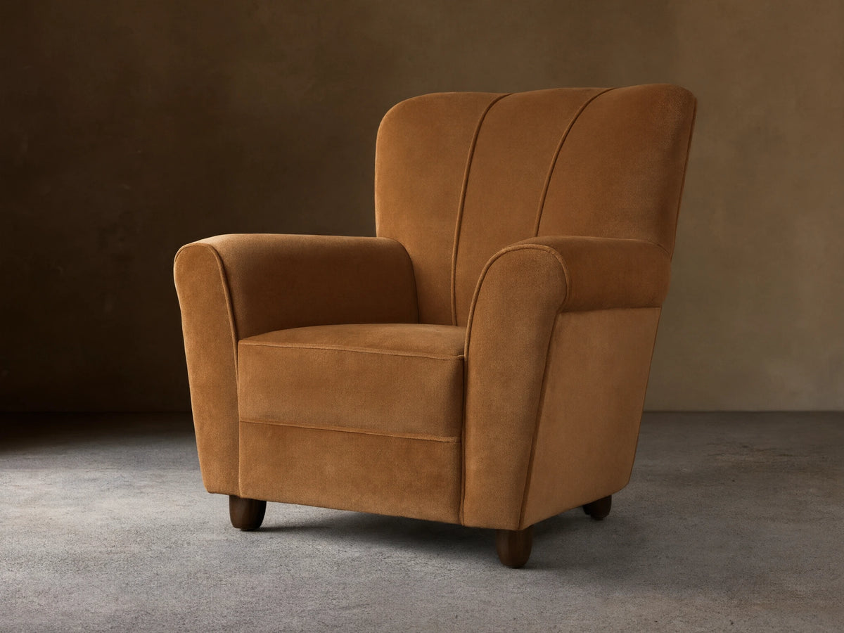 Prato Chair in Banks Camel velvet, angled. Features rolled arms, vertical channel stitching on back, on a wood floor.