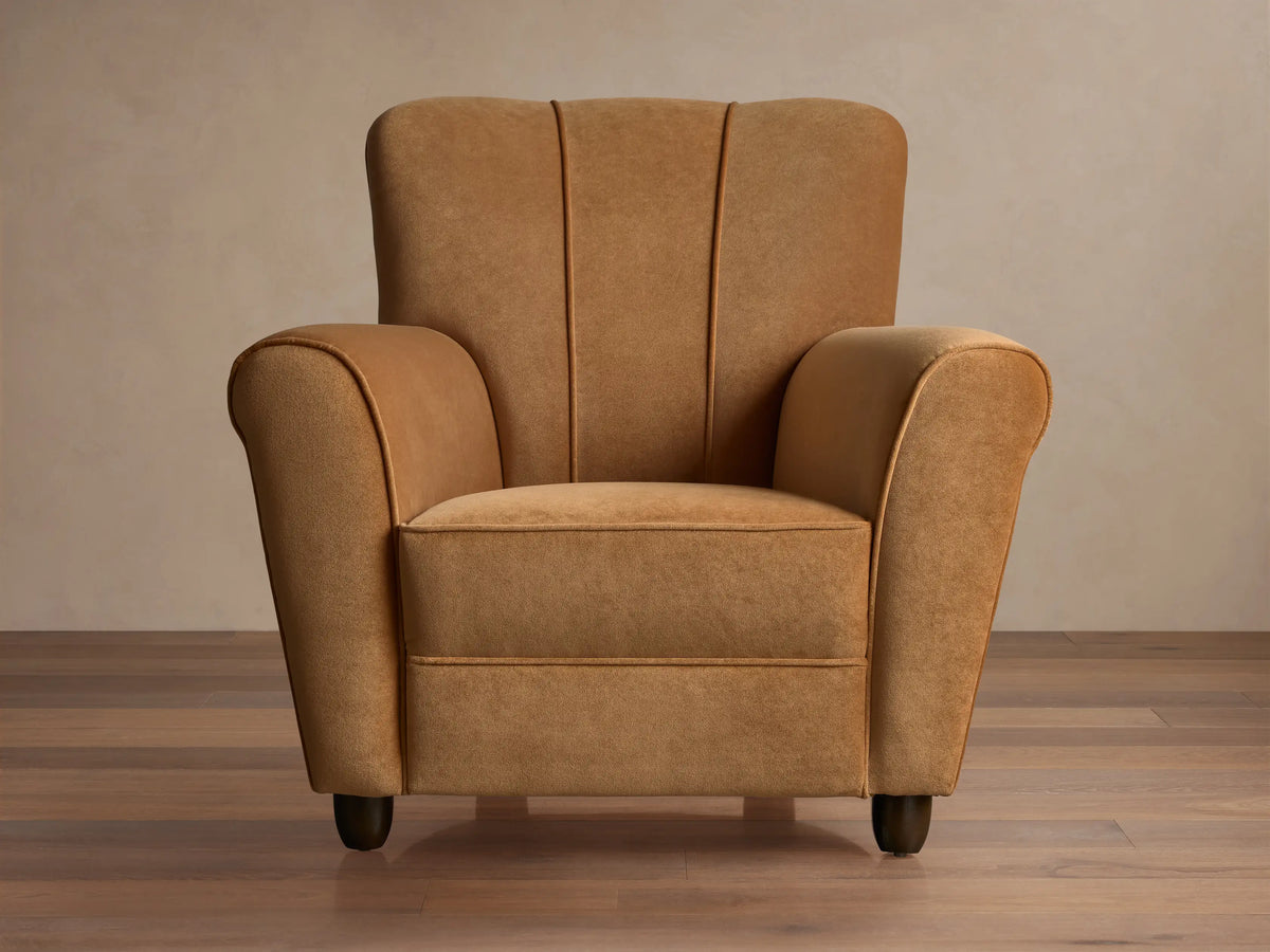 Prato Chair in Banks Camel velvet, angled. Features rolled arms, vertical channel stitching on back, on a wood floor.