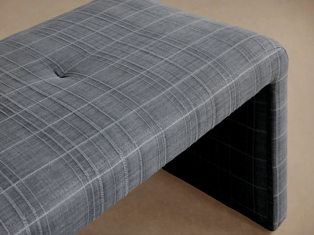 Ryan Bench: Dark gray plaid upholstered bench with a tufted top and waterfall design, against a beige wall on wood floor.