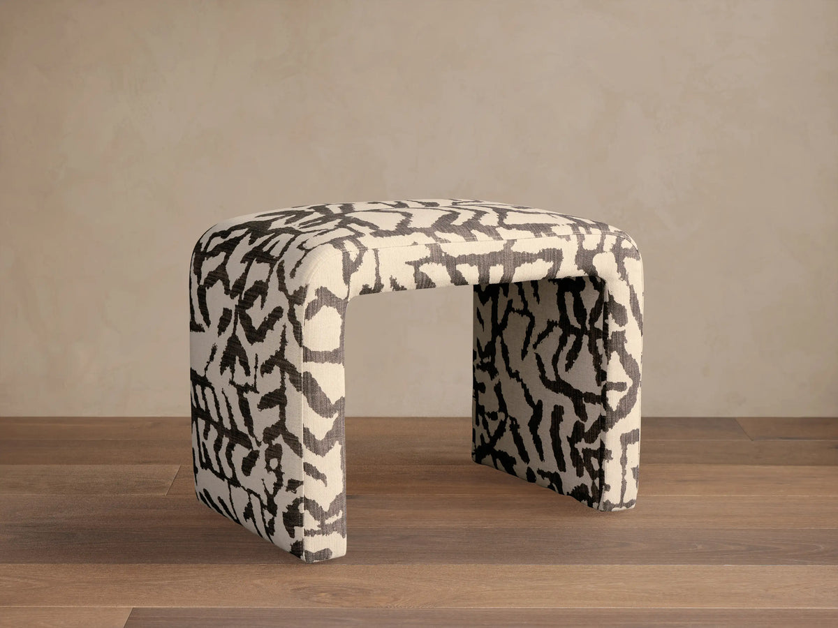 Ryan Petite Ottoman upholstered in black and off-white abstract patterned fabric, U-shaped, resting on a wooden floor, front view.