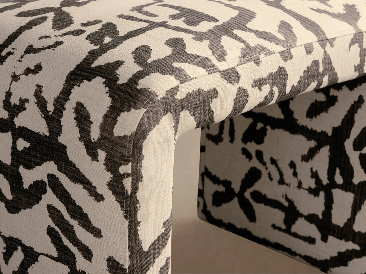 U-shaped Ryan Petite Ottoman upholstered in off-white fabric with dark gray abstract pattern, against a light wall.