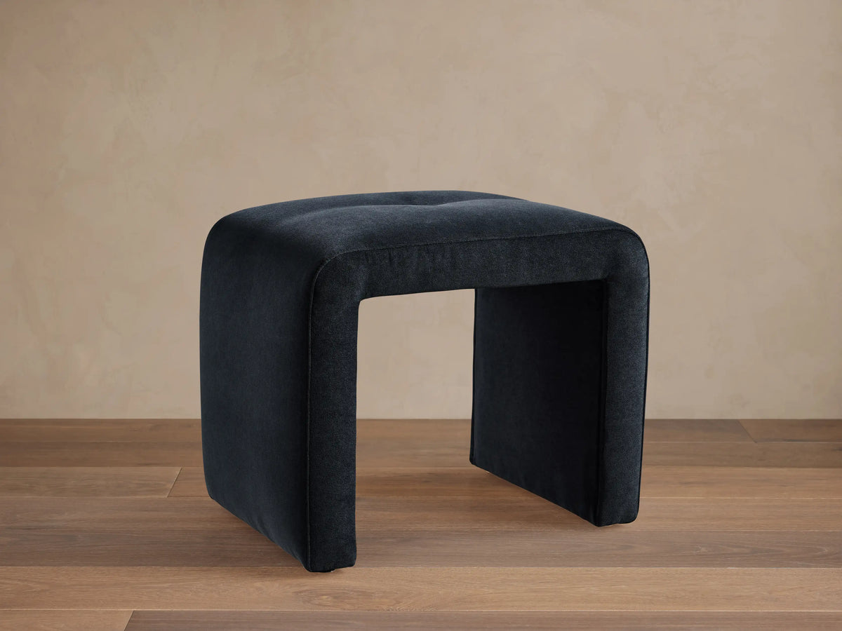 Ryan Mini Ottoman in dark blue, featuring a square shape with rounded edges, set on wooden floor and against a beige wall.