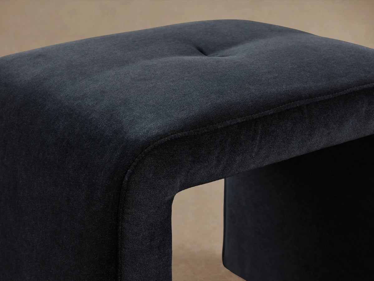 Ryan Mini Ottoman in dark fabric, featuring a simple cube-like shape with rounded edges, placed on a wooden floor against a beige wall.