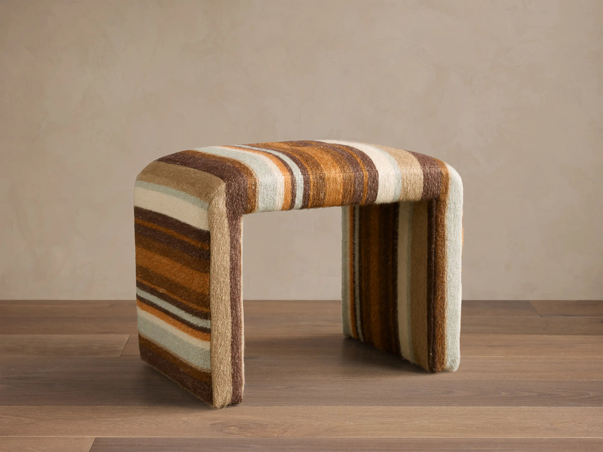 Front view of the Ryan Petite Ottoman with striped upholstery in shades of brown, orange, cream, and light blue. Rounded.