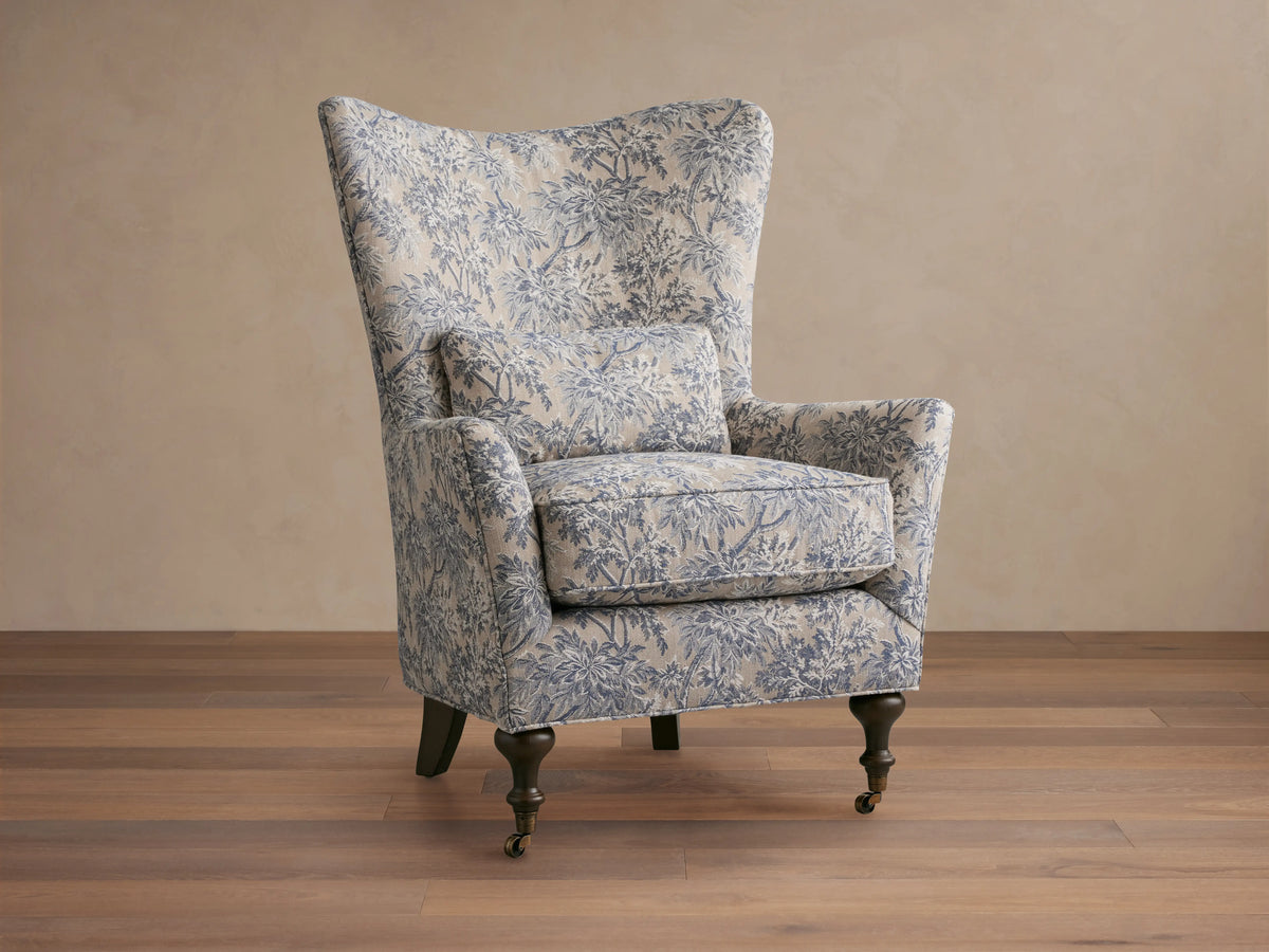Rio Chair with floral-patterned upholstery in soft blue tones, high wingback, plush seat cushion, solid wood legs, and brass caster wheels.