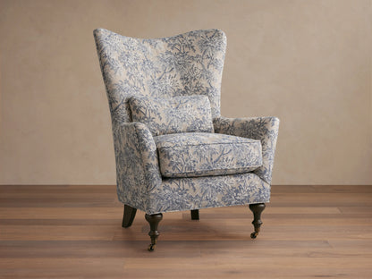 Rio Chair with floral-patterned upholstery in soft blue tones, high wingback, plush seat cushion, solid wood legs, and brass caster wheels.