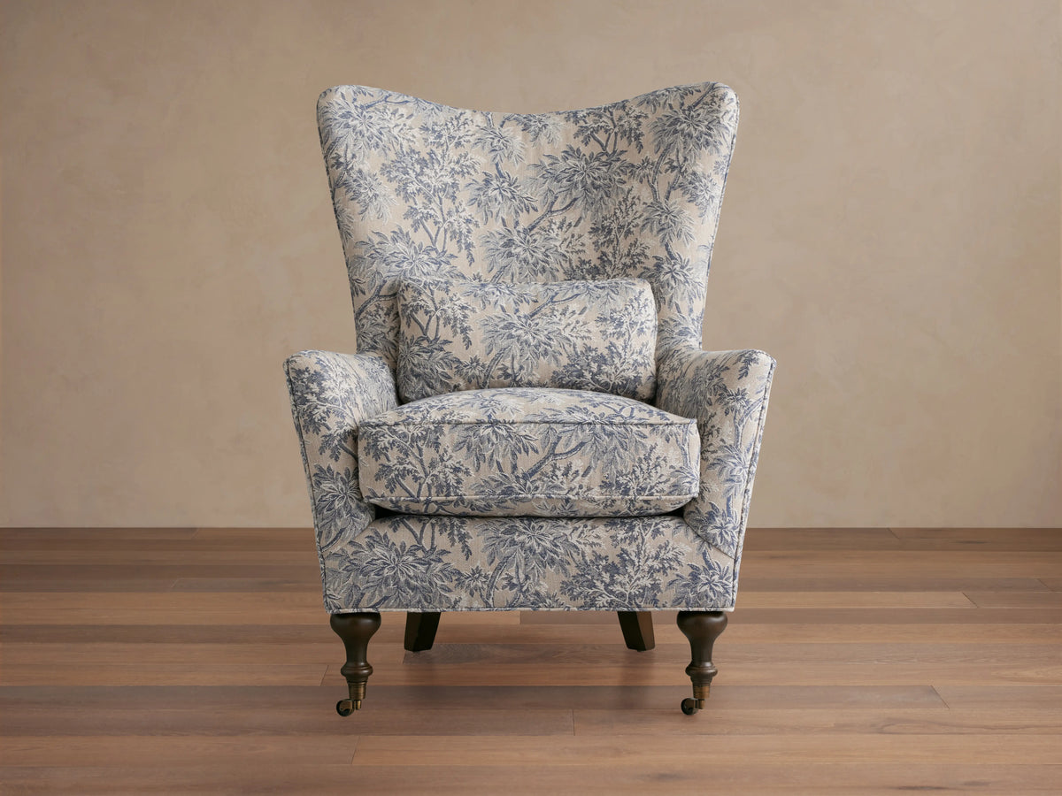 Rio Chair with floral-patterned upholstery in soft blue tones, high wingback, plush seat cushion, solid wood legs, and brass caster wheels.
