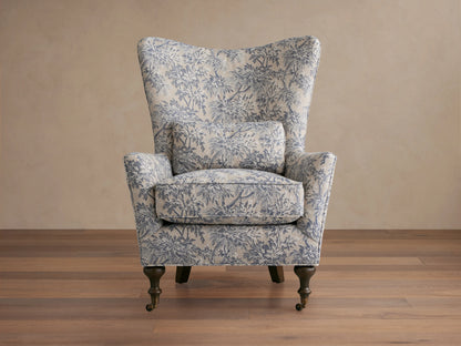 Rio Chair with floral-patterned upholstery in soft blue tones, high wingback, plush seat cushion, solid wood legs, and brass caster wheels.