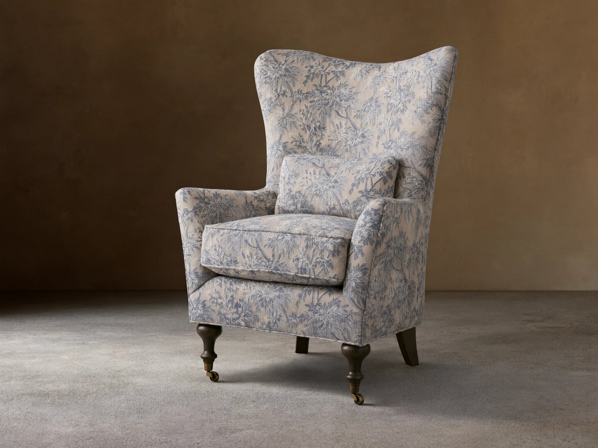 Rio Chair with floral-patterned upholstery in soft blue tones, high wingback, plush seat cushion, solid wood legs, and brass caster wheels.
