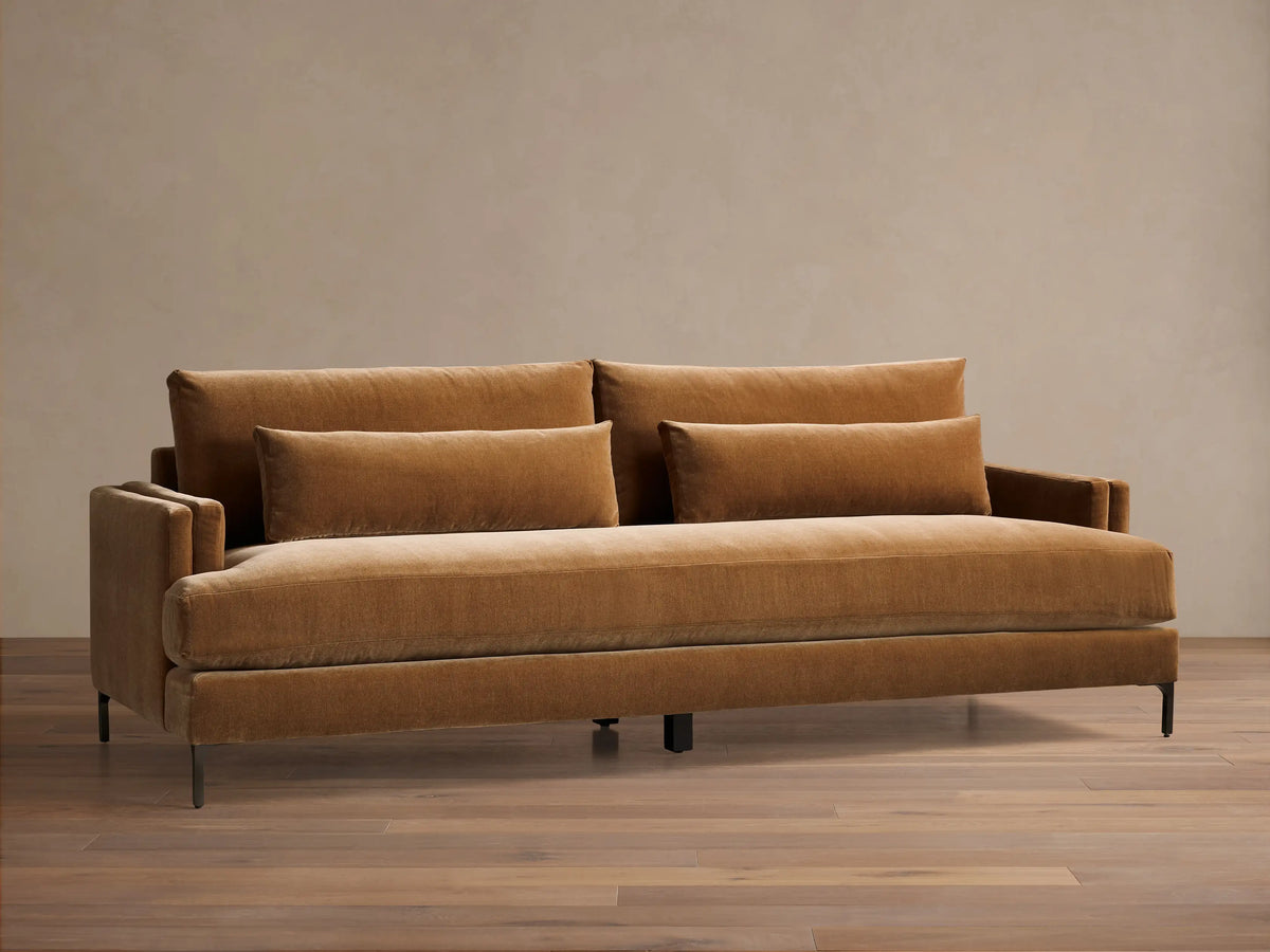 Front view of a caramel velvet sofa, featuring two back cushions, two throw pillows, and dark metal legs.
