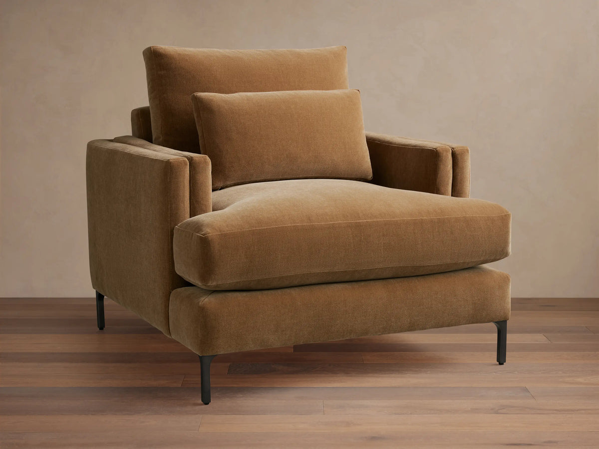Sumner Chair in brown fabric, showing plush seat and back cushions with supportive armrests, on slender black legs on wooden floor.