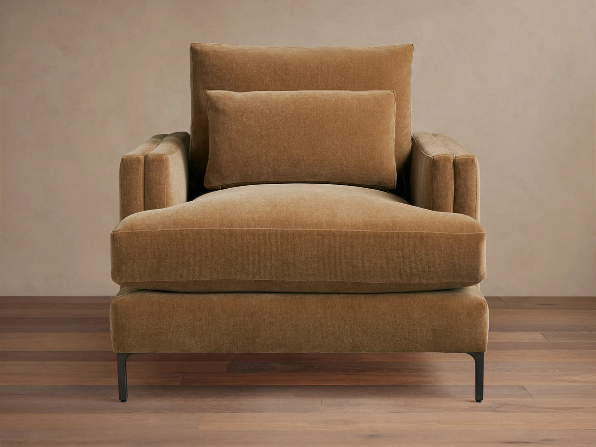 Sumner Chair in brown fabric, showing plush seat and back cushions with supportive armrests, on slender black legs on wooden floor.