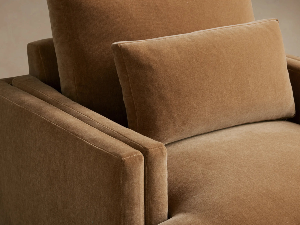 Sumner Chair in a warm brown fabric, viewed from the front. Features plush seat and back cushions, slim black legs on a wood floor.