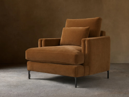 Sumner Chair in brown fabric, showing plush seat and back cushions with supportive armrests, on slender black legs on wooden floor.