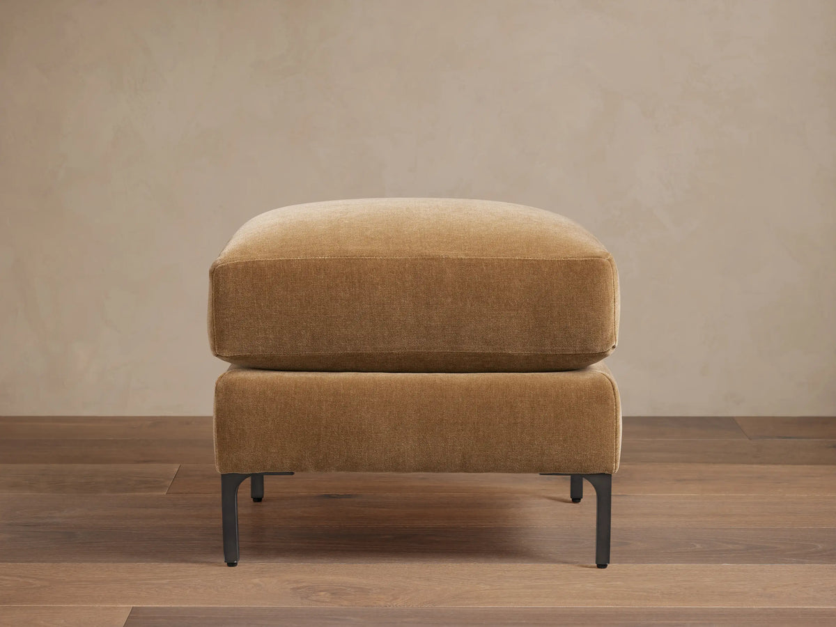 Sumner Ottoman in tan fabric on dark wooden floor, minimalist design, square shape, resting on slim black metal legs.