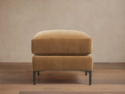Sumner Ottoman in tan fabric on dark wooden floor, minimalist design, square shape, resting on slim black metal legs.