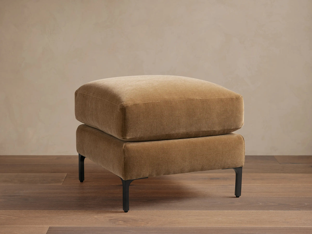 Sumner Ottoman in tan fabric on dark wooden floor, minimalist design, square shape, resting on slim black metal legs.
