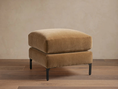 Sumner Ottoman in tan fabric on dark wooden floor, minimalist design, square shape, resting on slim black metal legs.