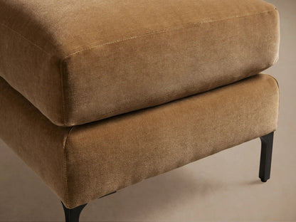 Sumner Ottoman upholstered in a light brown fabric, featuring a cushioned top and dark slim legs, set against a wooden floor and neutral wall.