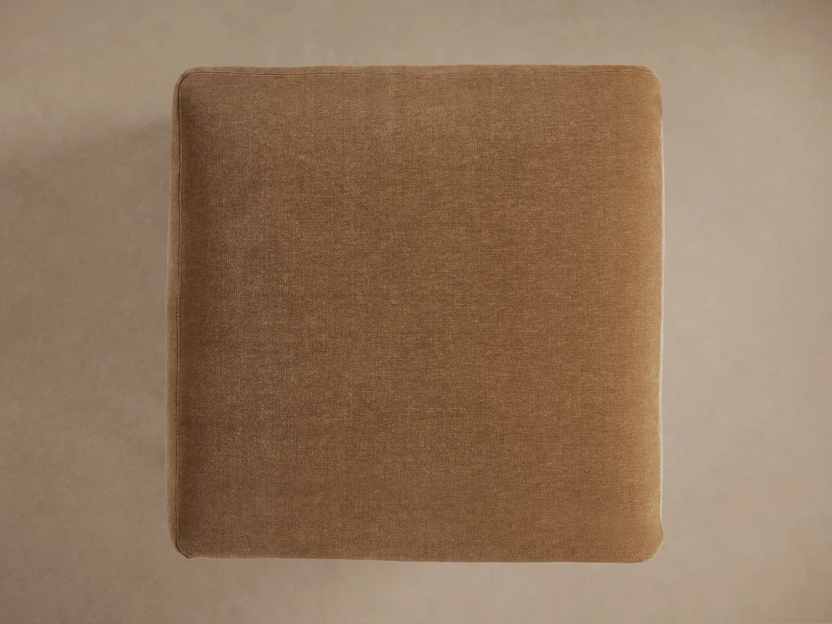 Sumner Ottoman in tan fabric, featuring soft, plush cushion with black legs, positioned on a neutral background.